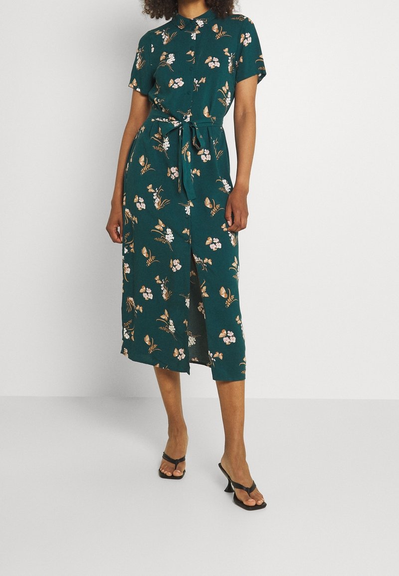 Femme portant une robe midi verte à fleurs avec manches courtes, ceinture à la taille et sandales à talons noires, debout devant un fond uni.
