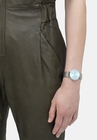 Zilveren metalen horloge met een lichtblauwe wijzerplaat en minimalistisch ontwerp, gedragen aan een hand boven de textuur van een olijfgroene leren overall.