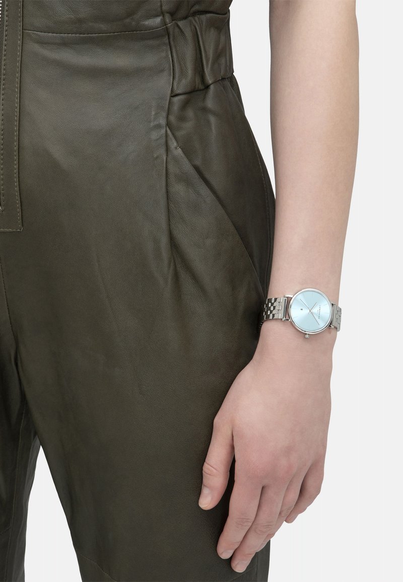 Zilveren metalen horloge met een lichtblauwe wijzerplaat en minimalistisch ontwerp, gedragen aan een hand boven de textuur van een olijfgroene leren overall.