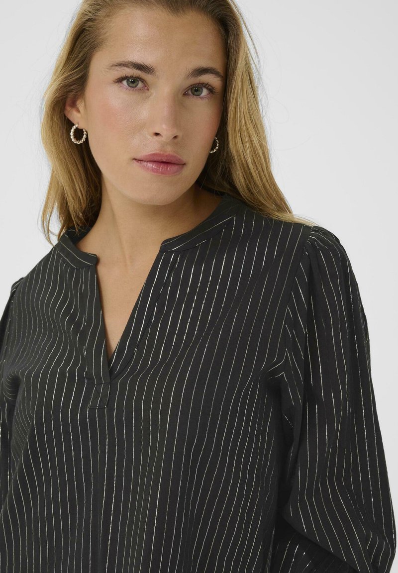 Jeune femme aux cheveux blonds portant des boucles d'oreilles créoles et une blouse noire avec de fines rayures blanches verticales.