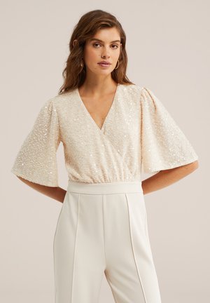Kvinde med bølget brunt hår iført en cremefarvet, palietbesat V-halsbluse med vide korte ærmer og cremefarvede bukser med høj talje mod en neutral baggrund.