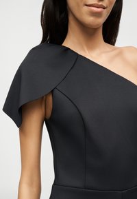 Γυναίκα που φοράει εφαρμοστό μαύρο φόρεμα με έναν ώμο και μεγάλο ασύμμετρο μανίκι στην αριστερή πλευρά, μπροστά σε απλό ανοιχτό φόντο.