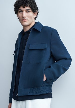 Joven con cabello rizado y oscuro, vestido con una chaqueta azul marino con bolsillos con solapa y dobladillo elástico, de pie frente a un fondo gris claro.