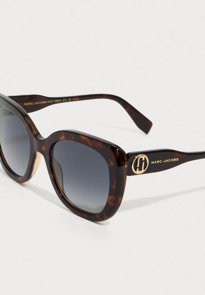 Gafas de sol en forma de ojo de gato en carey con lentes degradados oscuros, logotipo dorado de Marc Jacobs en las patillas y brazos curvados sobre fondo blanco.