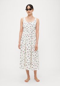 Balta gėlėta suknelė su margais mažais žiedeliais, be rankovių, laisvai krentanti, vidutinio ilgio sijonu ir su prigludusia juosmens dalimi.