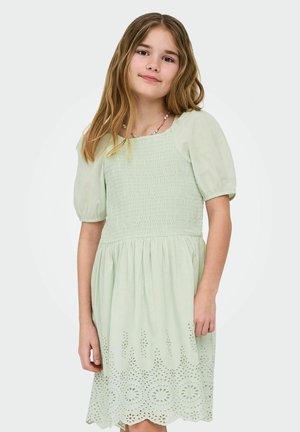 Jeune fille aux longs cheveux portant une robe vert clair à manches bouffantes et ornée de broderies anglaises à l'ourlet, sur un fond uni.