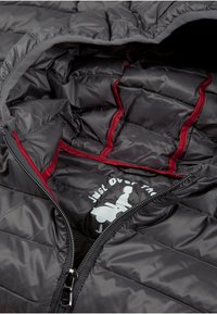 De binnenkant van de zwarte pufferjack laat een gequilte vormgeving zien met rode accenten en een wit logo. Het heeft een ritsluiting en een zachte, glanzende stofstructuur.