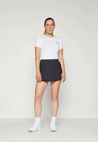 Λευκή κοντομάνικη εφαρμοστή μπλούζα συνδυασμένη με σκούρα αθλητική φούστα. Το μοντέλο φοράει λευκά αθλητικά παπούτσια με λευκές ψηλές κάλτσες. Απλό λογότυπο στην μπλούζα.