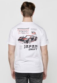 Balta trumpa rankove marškinėliai su grafiniu dizainu, vaizduojančiu sportinį automobilį, tekstu "TOKYO" ir "JAPAN DRIFT" ryškiais, išraiškingais rašmenimis.