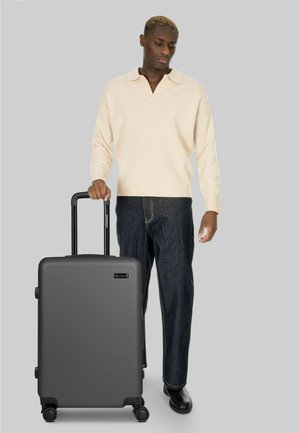 Hombre con suéter crema y vaqueros oscuros, de pie junto a una gran maleta gris con asa extendida sobre fondo gris.