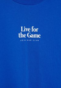 Chemise bleue avec texte blanc indiquant « Live for the Game » et un texte plus petit en dessous disant « Rejoignez notre club », centré sur la zone de la poitrine.