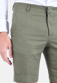 Bermuda in lino verde oliva con una texture liscia, dotate di tasche laterali e orlo arrotolato. Indossate con una camicia bianca a maniche lunghe.