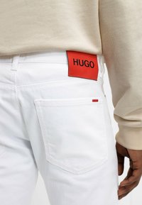 Weiße Jeans mit einem auffälligen roten "HUGO"-Label auf der Gesäßtasche, ausgestattet mit Standardtaschen und einer glatten Textur. Beige Oberteil sichtbar.