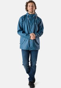 Blå vattentät jacka med huva, framknäppning och dragsko. Två sidofickor och en slät yta. Kombinationen med blå jeans och svarta skor.