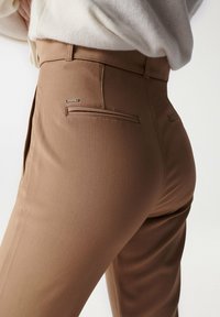 Brune skræddersyede bukser med en glat tekstur, der har en enkelt baglomme og metallisk detaljering. Taljen er tætsiddende for et struktureret look.