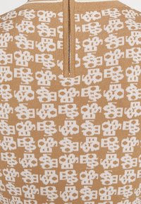 Pull en maille marron avec un motif blanc. Le design comprend des formes répétitives. Il possède une fermeture éclair au dos.