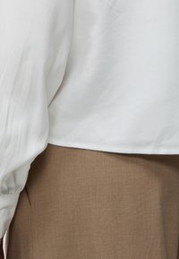 Blouse blanche à manches longues avec poignets froncés portée sur un pantalon ou une jupe marron taille haute.