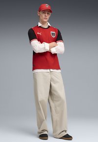 Κόκκινη και μαύρη αθλητική φανέλα με λογότυπο, φορεμένη πάνω από λευκό πουκάμισο. Μπεζ φαρδιές παντελόνες και  μαύρα παπούτσια με καφέ σόλες. Κόκκινο καπέλο με λογότυπο.