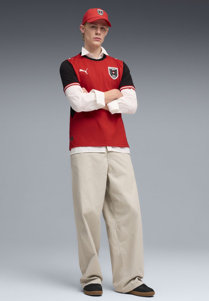 Κόκκινη και μαύρη αθλητική φανέλα με λογότυπο, φορεμένη πάνω από λευκό πουκάμισο. Μπεζ φαρδιές παντελόνες και μαύρα παπούτσια με καφέ σόλες. Κόκκινο καπέλο με λογότυπο.