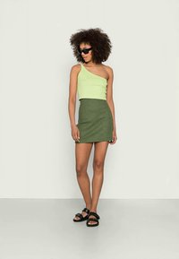 Mulher com cabelo encaracolado, usando óculos de sol pretos, blusa de um ombro verde-lima, saia verde xadrez e sandálias pretas, de pé contra uma parede branca.