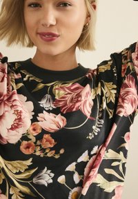Černý top s květinovým vzorem v odstínech růžové, žluté a bílé. Hribkový výstřih s kulturními zlatými akcenty. Měkká, texturovaná tkanina.