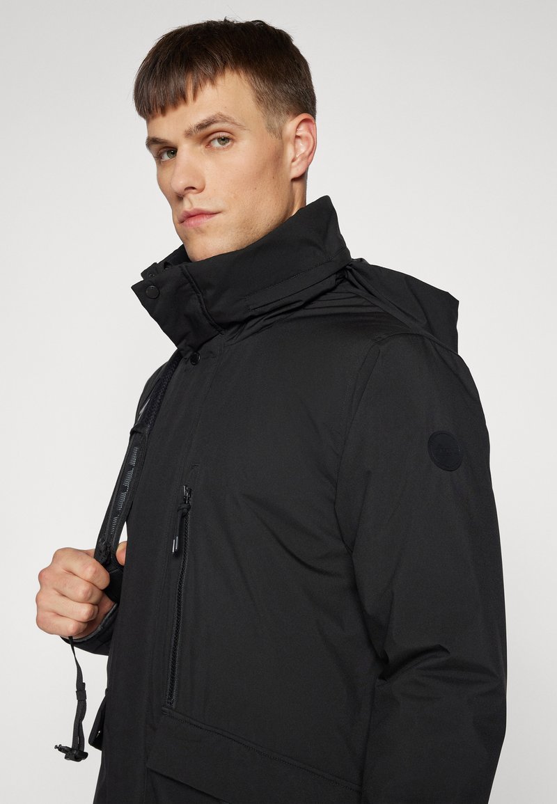 Czarna pikowana kurtka z wysokim kołnierzem, kieszeniami zapinanymi na zamek oraz regulowanym ściągaczem. Gładka tekstura o matowym wykończeniu.