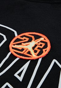 Oransje og gul brodert rund lapp med silhuett av en basketballspiller som hopper og stilisert labyrintmønster på svart stoff.