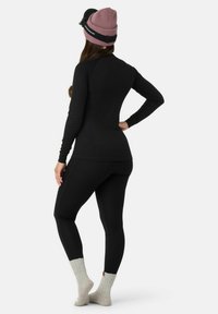 Schwarzes Thermaloberteil und Leggings aus geripptem Material, kombiniert mit einer hellrosanen Strickmütze und grauen Socken mit umgeschlagenem Bündchen, von hinten betrachtet.