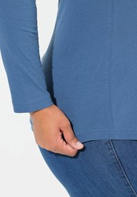 Lange mouw blauwe katoenen top, met een ronde halslijn en vlakke naden, gecombineerd met donkerblauwe denim. Close-up van een hand bij de zoom.