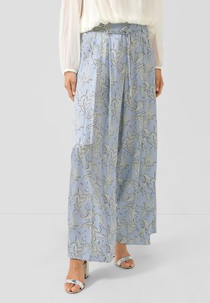 Frau trägt hellblaue weite Hosen mit weißem Blumenmuster und silbernen Sandalen mit Absatz, steht vor einem schlichten grauen Hintergrund.