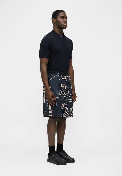 Άνδρας όρθιος πλευρικά, φοράει μαύρο πόλο μπλουζάκι με κοντά μανίκια, σορτς μέχρι το γόνατο με σχέδιο, μαύρες κάλτσες και μαύρα αθλητικά παπούτσια σε απλό φόντο.