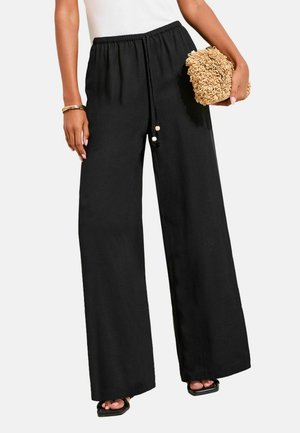 Frau trägt schwarze Hosen mit weitem Bein und Kordelzug am Bund, hält eine strukturierte beige Clutch, trägt schwarze Sandalen und goldene Armbänder.