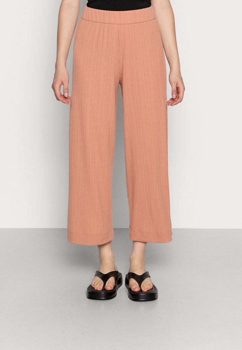 Femme portant un pantalon pêche taille haute, jambes larges et coupé au-dessus de la cheville avec des rayures verticales, ainsi que des sandales à plateforme noires, sur un fond uni.