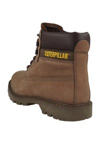 Botín de cuero marrón hasta el tobillo con una textura suave, suela de goma, ojales de metal y logo amarillo de "Caterpillar" en el lateral. Diseño de punta redonda.