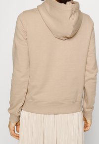 Béžová mikina s kapucí, měkkou texturou, žebrovanými manžetami a pásovým lemem. Má jednoduchý design, který zdůrazňuje čistý a minimalistický vzhled.