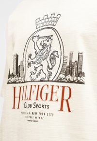 Έμβλημα λιονταριού με κορωνίδα πάνω από τον ορίζοντα της πόλης, μεγάλο κείμενο "HILFIGER" και μικρότερο "Club Sports Manhattan New York City Columbus Avenue" σε μπεζ ύφασμα.