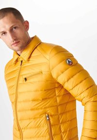 Geel pufferjack heeft horizontale quilting, een rits aan de voorkant en een borstzak. Inclusief een logobadge op de linker mouw. Luxe textuur.