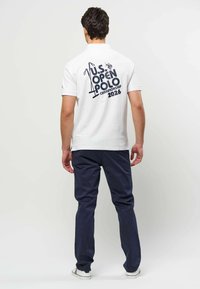 Homme portant un polo blanc avec un texte et des graphismes bleu marine "U.S. Open Polo Championship 2026" dans le dos, un pantalon bleu marine et des baskets blanches.