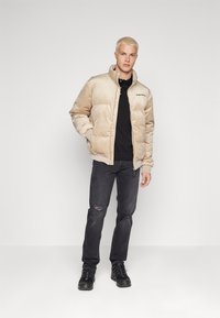 Giacca imbottita beige con colletto alto, zip frontale e logo. Indossata sopra una maglietta nera con jeans neri strappati e scarpe nere.