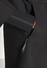 Veste imperméable noire à texture lisse. Elle comporte une poche avec un accent en tissu et une étiquette logo visible sur le côté.