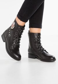 Mustat nahkaiset nilkkasaappaat, jossa on nauhakiinnitys, vetoketjukiinnitys ja koristeellisia patch-painatuksia. Matala lohkoheel ja teksturoidut mustat nauhat. Litteä pohja.