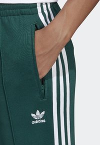 Zielone spodnie sportowe z białymi paskami po bokach i logo, z bocznymi kieszeniami i miękkim, teksturowanym materiałem. Zbliżenie na dłoń w kieszeni.