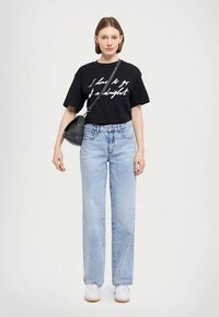 Svart bomulls-T-shirt med vit text, ljusblå högmidjade jeans i denim och vita sneakers; modellen håller en liten svart handväska.