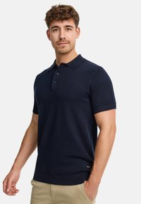 Polo à manches courtes bleu marine en tricot texturé, avec un col, trois boutons et un ourlet côtelé. Le modèle porte un pantalon beige.