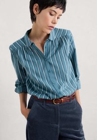 Turkusowa koszula z guzikami, w białe pionowe paski, z miękkim kołnierzykiem i zgromadzonymi ramionami, w połączeniu z granatowymi spodniami z kordurowej tkaniny i plecionym brązowym pasem.