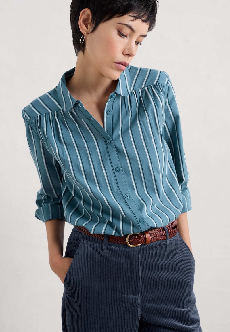Turkusowa koszula z guzikami, w białe pionowe paski, z miękkim kołnierzykiem i zgromadzonymi ramionami, w połączeniu z granatowymi spodniami z kordurowej tkaniny i plecionym brązowym pasem.