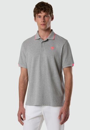 Uomo che indossa una polo grigia con rifiniture e logo rosa, abbinata a pantaloni bianchi, in piedi davanti a uno sfondo chiaro e semplice.