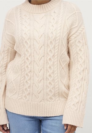 Cream-colored cable knit sweater with ribbed hem and cuffs worn with light blue jeans, focusing on texture and knit patterns.