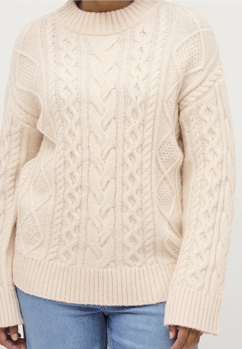 Pull en maille torsadée couleur crème avec ourlet et poignets côtelés porté avec un jean bleu clair, mettant l'accent sur la texture et les motifs de tricot.