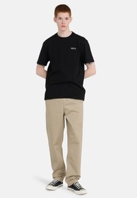 Sort bomulds T-shirt med korte ærmer og et lille logo på brystet, parret med beige bukser og sorte sneakers.